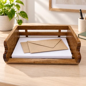 May include: A rectangular wooden document tray with a slatted design and a warm brown finish. The tray holds a stack of white paper sheets topped with two light brown kraft paper envelopes, providing an organized storage solution for desk items.