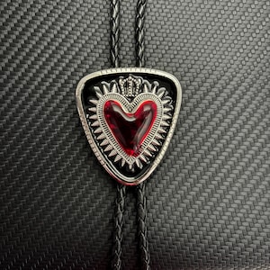 Può includere: Una cravatta bolo con un ciondolo triangolare in metallo argentato e un cuore rosso al centro. Il cuore è circondato da una cornice decorativa in argento con una piccola corona sulla parte superiore. Il ciondolo è fissato a un cordino in pelle intrecciata.