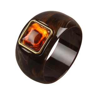May include: A wide, dark brown marbled resin bangle bracelet featuring a prominent, square-shaped amber-colored stone set within a polished gold-tone metal frame. The smooth, glossy surface of the resin displays swirling patterns of lighter brown.