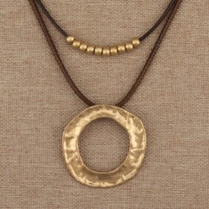 May include: A double-strand necklace featuring a dark brown cord. The upper strand is adorned with a row of nine small, matte gold-toned spherical beads. The lower strand holds a large, circular pendant with a textured, hammered gold-toned finish.