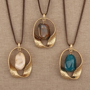 Puede incluir: Tres collares con cordones marrones y colgantes de metal dorado. Cada colgante consiste en un marco ovalado abierto, texturizado y ondulado que sostiene una piedra pulida, lisa e irregular en colores crema, marrón o azul cerceta.