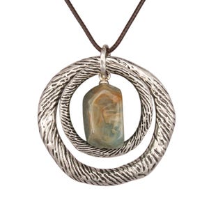 May include: A pendant necklace featuring two interlocking silver-toned rings with a textured, organic surface. A smooth, polished, multi-colored stone with swirls of tan, blue, and grey is suspended from the center of the rings on a dark brown cord.