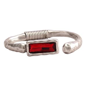 May include: An open-ended cuff bracelet crafted from silver-toned metal with a hammered texture. The design features a rectangular, deep red gemstone set in a frame, positioned next to a section of coiled wire detailing on the band.
