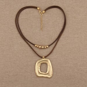 Puede incluir: Un collar de doble hebra con cordón marrón oscuro y un colgante grande de color dorado mate con forma irregular y una abertura central. El collar incluye cinco pequeñas cuentas redondas de color dorado mate en la hebra superior y una cadena extensora.