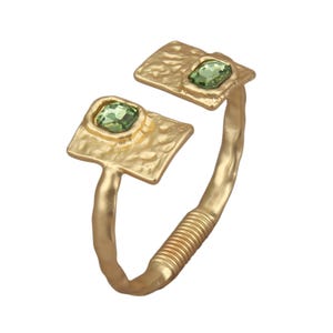 May include: A gold-coloured cuff bracelet with a textured surface. The bracelet has two square settings, each holding a green gemstone. The band features a wrapped wire design near the opening, creating a unique and stylish accessory.
