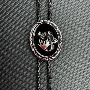 May include: A silver-toned bolo tie with a black oval pendant. The pendant features a silver heart with flames and an arrow design. The braided black cord completes the western-inspired accessory.