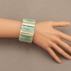 May include: A wide bracelet with rectangular segments in shades of green and gold. The bracelet is worn on a wrist, showcasing its design and color contrast. The bracelet's segments are approximately 1 inch wide.