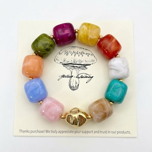 May include: A colorful bracelet featuring square beads in various colors, including pink, blue, green, and yellow. The bracelet has gold-colored accents and is displayed on a card with a mushroom illustration and the text "Jelly Lainey".