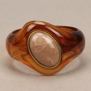 May include: A wide bangle bracelet made of translucent amber-colored resin with a swirling pattern. The center features a large, smooth, oval-shaped beige cabochon stone set within a thin, polished gold-tone metal frame.