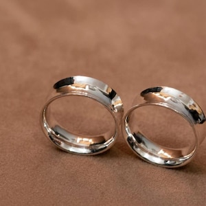May include: Two polished silver-toned metal rings with a smooth, rounded exterior surface and a reflective finish. The bands feature a consistent width and a clean, circular design, showcasing a bright metallic luster that catches the light.