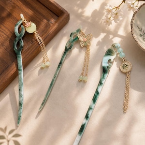May include: Three jade hair sticks with intricate designs. The hair sticks are green and white, with gold-colored chain accents and decorative charms. One stick features a butterfly, another a round charm, and the third a floral design.