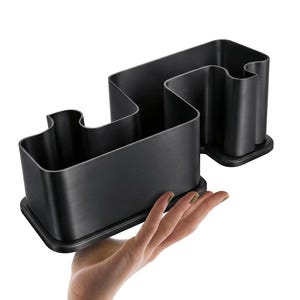 Op de afbeelding: Een zwarte bureau-organizer met een uniek, golfachtig ontwerp. De organizer heeft meerdere compartimenten van verschillende afmetingen en staat op een rechthoekige basis. Het product wordt in een hand gehouden, wat de grootte en vorm laat zien.