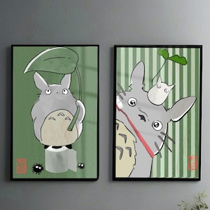 May include: Two framed art prints featuring the animated character Totoro. The left print shows Totoro holding a leaf umbrella against a green background. The right print depicts Totoro with a small white creature on its head, set against a striped green background.