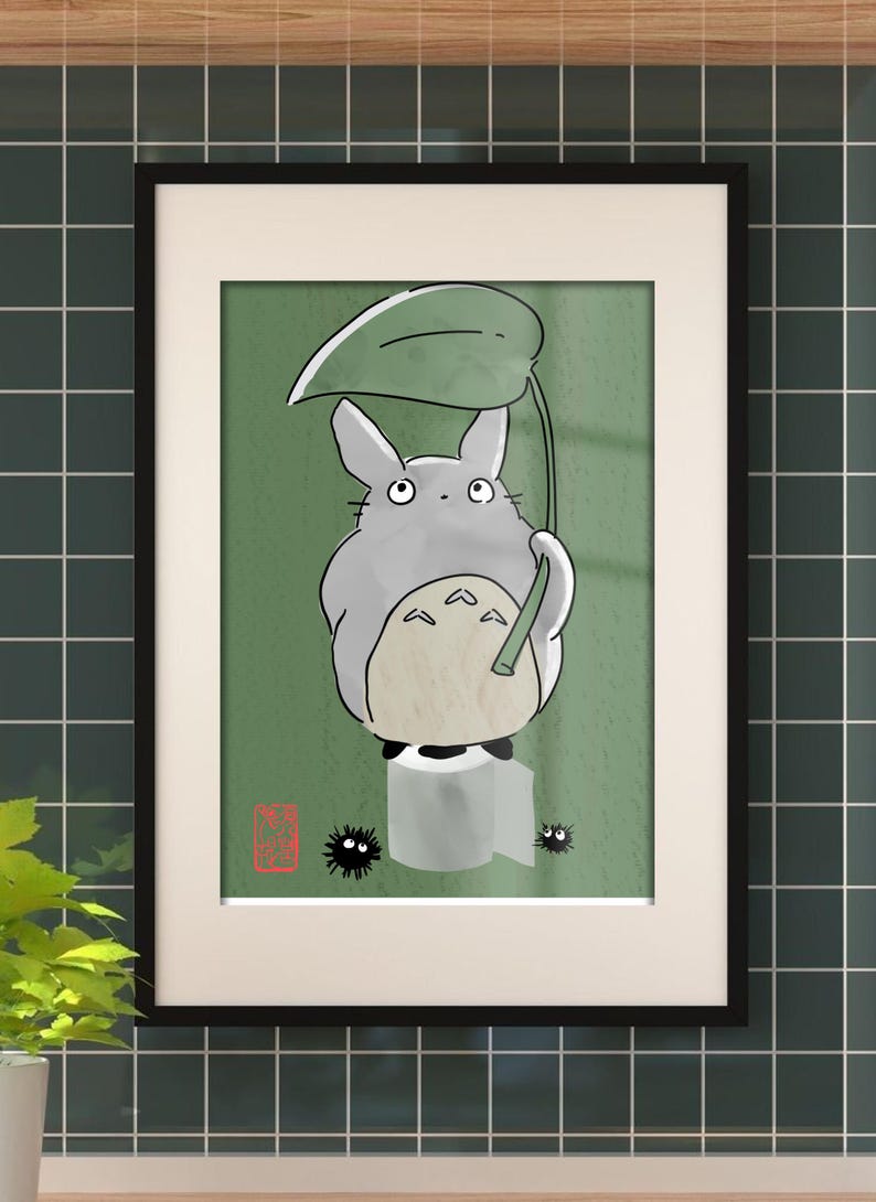 May include: Framed art print depicting a cartoon Totoro character with a leaf umbrella. The gray Totoro is sat on a roll of toilet paper against a green backdrop. Two small black soot sprites are also present. The print is in a black frame.
