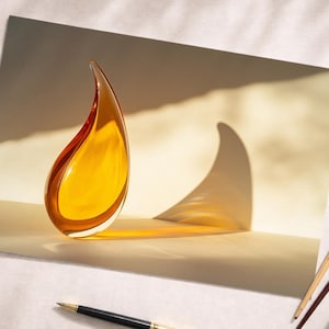 Könnte beinhalten: Eine tropfenförmige Skulptur aus bernsteinfarbenem Glas mit geschwungenem Design. Die Skulptur wirft einen Schatten auf eine helle Oberfläche. Ein Stift, zwei Pinsel und eine Kaffeetasse sind ebenfalls zu sehen.