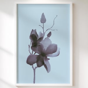 May include: A framed floral print featuring a detailed illustration of a magnolia flower in shades of purple and gray. The artwork is set against a light blue background within a white frame, creating a serene aesthetic.