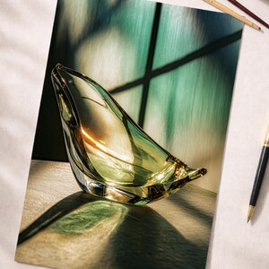 Könnte beinhalten: Eine klare, grüne Glasskulptur mit einer einzigartigen, asymmetrischen Form. Die Skulptur steht auf einer hellen Oberfläche, auf die Schatten geworfen werden. Ein Stift, Pinsel und eine Tasse Kaffee sind ebenfalls zu sehen.