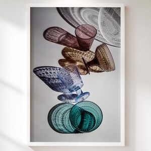 Könnte beinhalten: Ein gerahmter Druck mit einer künstlerischen Anordnung von farbigem Glasgeschirr. Das Bild zeigt verschiedene Gläser und Teller in Blau-, Grün-, Braun- und Klartönen, wodurch eine visuell ansprechende Komposition entsteht. Das Glasgeschirr ist in einem vertikalen Stapel angeordnet.