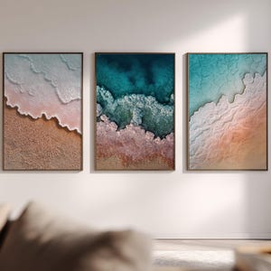 Könnte beinhalten: Drei gerahmte Wandkunstwerke, die Luftaufnahmen von Ozeanwellen zeigen, die auf das Ufer treffen. Die Kunstwerke zeigen eine Farbpalette aus Blau-, Rosa- und Brauntönen mit strukturierten Details. Jedes Stück ist rechteckig und dunkelbraun gerahmt.