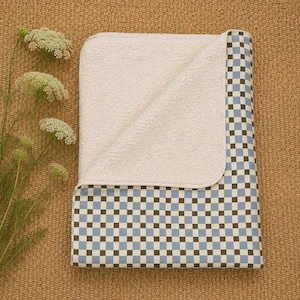 May include: A folded blanket featuring a light blue and dark brown checkered pattern on one side and a textured, cream-colored fleece lining on the reverse. The blanket is rectangular with rounded corners and a soft, plush appearance.