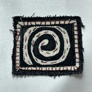 May include: A square fabric patch featuring a black background with a white spiral design. The patch has a frayed edge and is outlined with a decorative border of white fabric stitched with thin, dark brown thread throughout the design.