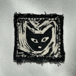 May include: A square fabric patch with a black and white graphic print of a cat face surrounded by abstract lines. The patch features a frayed, raw edge border and a high-contrast design, showcasing a stylised feline head centred within the square.