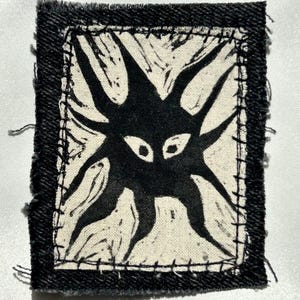 May include: A rectangular fabric patch featuring a black, multi-limbed creature graphic printed on an off-white background. The central design is framed by a thick black border with visible, coarse black stitching along the edges of the fabric.