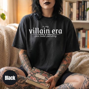 May include: Black t-shirt with the text "in my villain era plot twist pending" in white. The shirt is a dark charcoal color and has a relaxed fit. The text is centered on the front of the shirt.