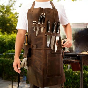 Könnte beinhalten: Eine braune Lederschürze mit mehreren Taschen und Messerhaltern. Die Schürze hat einen Flaschenhalter und einen Flaschenöffner. Mehrere Messer sind in den Messerhaltern verstaut. Die Schürze wird von einer Person in einem weißen T-Shirt getragen.