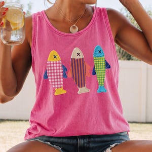 May include: A bright pink sleeveless tank top featuring three embroidered fish appliqués on the front. The fish are decorated with colorful patterns including pink gingham, orange and purple stripes, and a green and yellow checkered design.