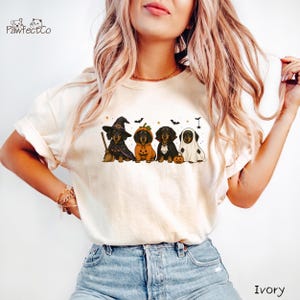 May include: An ivory t-shirt featuring a graphic of four dachshunds dressed in Halloween costumes, including a witch, a pumpkin, a vampire, and a ghost, with small bats and stars surrounding them. The text PawfectCo is printed in the top left corner.