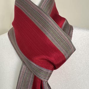 May include: A scarf featuring a deep red solid center panel flanked by wide borders with a fine, multi-colored pinstripe pattern in shades of green, grey, and purple. The fabric has a subtle sheen and is draped around a white mannequin neck.