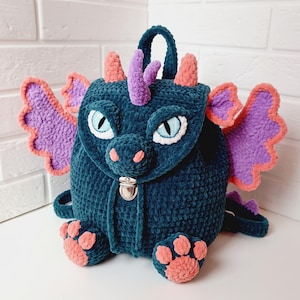 May include: A dark teal crochet backpack shaped like a dragon with purple wings, peach-colored horns, and peach-colored paw pads. The bag features light blue eyes, a silver metal clasp closure, and a top handle, crafted from soft plush yarn.