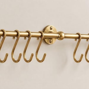 May include: A horizontal brass-toned metal rail featuring three circular wall-mounting plates with visible screws. The rail includes eight removable S-shaped hooks suspended from the rod, designed for hanging various items in a linear arrangement.