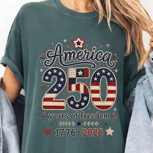May include: A blue spruce t-shirt with the text "America 250" in a patriotic design. The number 250 is filled with a stars and stripes pattern. The shirt also includes the text "years of freedom" and the years "1776-2026".