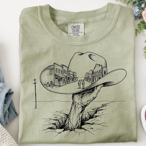 May include: Sage green Comfort Colors t-shirt featuring a black and white graphic. The graphic depicts a hand emerging from a crack in the ground, holding a cowboy hat with a town scene inside. The shirt is folded.
