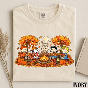 May include: An ivory t-shirt featuring a graphic print of the Peanuts characters sitting around a campfire in an autumn forest. The design includes Snoopy, Charlie Brown, and friends surrounded by orange leaves, pumpkins, and string lights.
