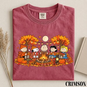 May include: A maroon crew neck t-shirt featuring a graphic of the Peanuts characters sitting around a campfire in an autumn forest. The design includes Snoopy, Charlie Brown, and friends with pumpkins, a lantern, and string lights under a full moon.