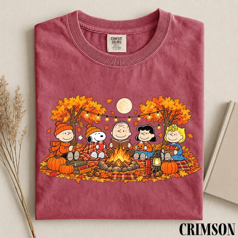 May include: A maroon crew neck t-shirt featuring a graphic of the Peanuts characters sitting around a campfire in an autumn forest. The design includes Snoopy, Charlie Brown, and friends with pumpkins, a lantern, and string lights under a full moon.