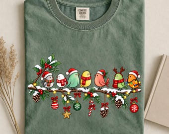 May include: A sage green t-shirt featuring a graphic of seven colorful birds perched on a snowy branch decorated with Christmas ornaments, pinecones, and holly. The neck label reads Comfort Colors. The design includes red and white accents.