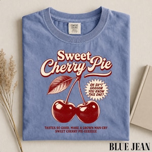 May include: A blue crew neck t-shirt featuring a graphic of two red cherries with a leaf. The text reads: Sweet Cherry Pie, Oh Sh*t Graham You Know This One?, Tastes So Good, Make A Grown Man Cry Sweet Cherry Pie-Eeeeeee. The shirt has a Comfort Colors label.
