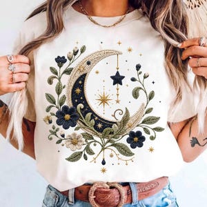 Op de afbeelding: Crèmekleurig T-shirt met een hemels ontwerp. Een halve maan, versierd met gouden borduursels en marineblauwe accenten, is omgeven door bloemen- en stermotieven. Het ontwerp bevat een hangende ster en een gouden ketting.