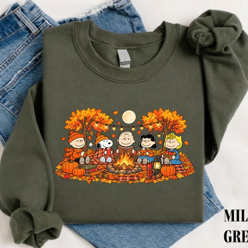 May include: An olive green crewneck sweatshirt featuring a graphic print of the Peanuts characters sitting around a campfire in an autumn forest. The scene includes pumpkins, fall foliage, string lights, and a full moon, all rendered in warm colors.