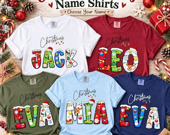 May include: Five short-sleeved t-shirts in white, maroon, olive green, light blue, and navy blue. Each shirt features the text Merry Christmas above a name spelled in large, colorful letters decorated with holiday ornaments and snow.