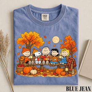 May include: A blue t-shirt featuring a graphic of the Peanuts characters sitting on a wooden bench under autumn trees. The design includes Snoopy, Charlie Brown, and friends holding mugs with pumpkins and fallen leaves on the ground below.