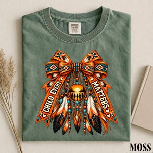 May include: A sage green t-shirt featuring a graphic of a large orange bow with geometric patterns, dangling feathers, and beads. In the center is a circular sunset scene with silhouettes of children. The text reads CHILD EVERY MATTERS.