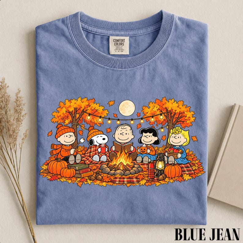 May include: A blue crew neck t-shirt featuring a graphic print of the Peanuts characters sitting around a campfire in an autumn forest. The design includes Snoopy, Charlie Brown, and friends with pumpkins, a lantern, and string lights.