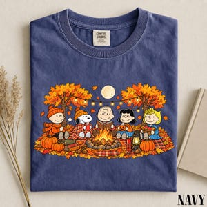 May include: A navy blue short-sleeve t-shirt featuring a graphic of the Peanuts characters sitting around a campfire in an autumn forest. The design includes Snoopy, Charlie Brown, and friends with pumpkins, a lantern, and string lights.