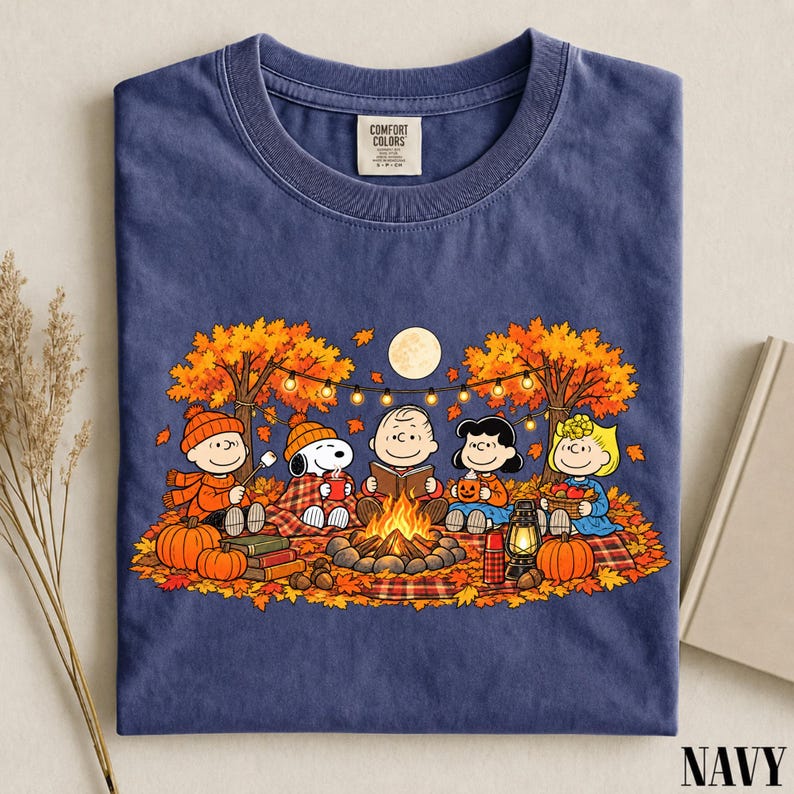 May include: A navy blue short-sleeve t-shirt featuring a graphic of the Peanuts characters sitting around a campfire in an autumn forest. The design includes Snoopy, Charlie Brown, and friends with pumpkins, a lantern, and string lights.