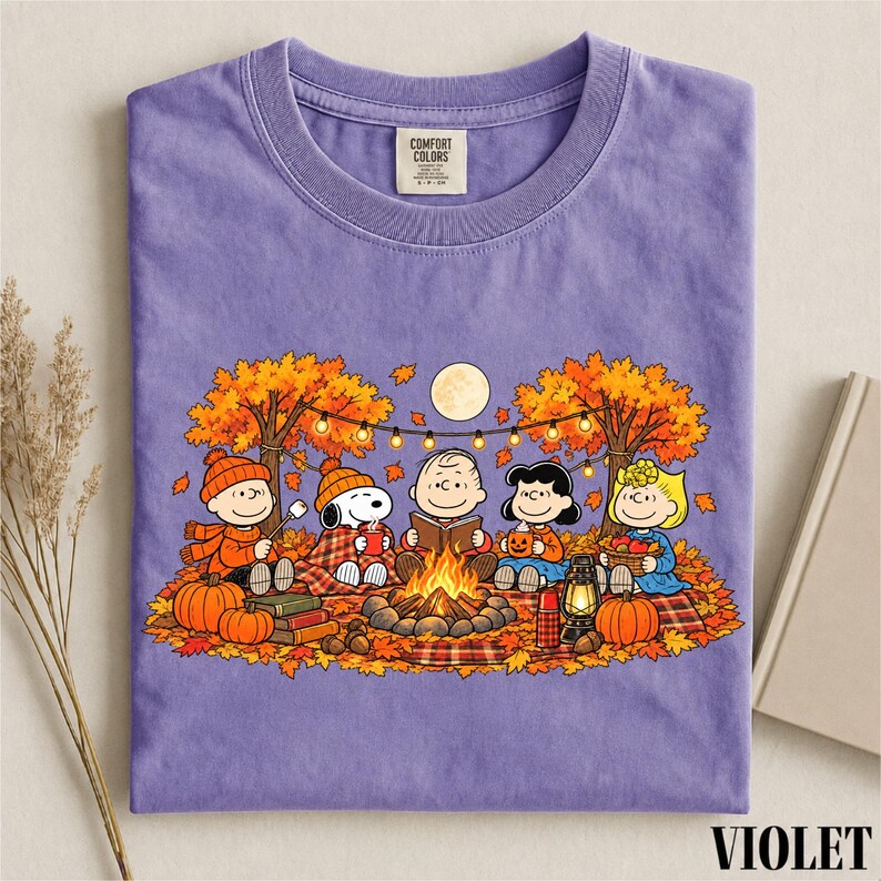 May include: A purple crew neck t-shirt featuring a graphic of the Peanuts characters sitting around a campfire in an autumn forest. The design includes Snoopy, Charlie Brown, and friends with pumpkins, a lantern, and string lights under a full moon.